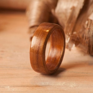 May include: A close-up of a wooden ring with a smooth, polished finish. The ring is a rich brown color and has a natural wood grain pattern. The ring is lying on a wooden surface.