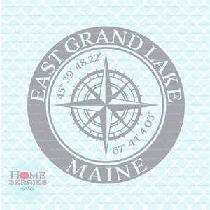 Op de afbeelding: Een grijs kompas met een blauwe achtergrond en de tekst "East Grand Lake, Maine" en coördinaten "45° 39' 48.22" en "67° 44' 4.03".