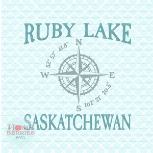 May include: A teal blue compass rose with the text "RUBY LAKE" and "SASKATCHEWAN" in a teal blue font. The compass rose has the coordinates "52°57' 41.8" W" and "102° 21' 20.5" E" written around it.