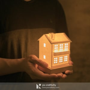 May include: A small, white paper house with windows and a balcony, lit from within, held in a person's hands.