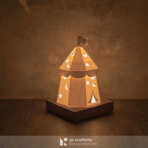 May include: A papercraft nightlight shaped like a tent with a flag on top. The tent has cutouts of stars and moons. The nightlight is lit up and casting a warm glow.