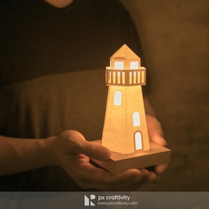 May include: A papercraft lighthouse with a light inside, held in a person's hands. The lighthouse is made of brown paper and has a white base.