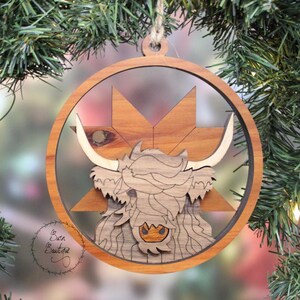 May include: A wooden Christmas ornament featuring a Highland cow with a brown and white patterned design. The ornament is shaped like a circle with a starburst design in the background.