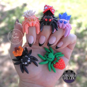 Puede incluir: Un conjunto de seis anillos de araña coloridos hechos de arcilla polimérica. Las araignées llevan varios accesorios, incluyendo una flor, un hongo, una fresa y un lazo. Los anillos se muestran en una mano con esmalte de uñas negro y blanco.