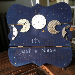 Puede incluir: Un letrero de madera azul con purpurina dorada y el texto "it's just a phase" escrito en blanco. El letrero está decorado con cuatro piezas de reloj antiguas que se asemejan a las fases de la luna.