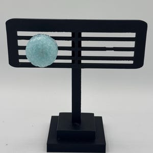 May include: A light blue glass orb with a textured surface is displayed on a black metal stand with a square base.