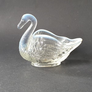 Puede incluir: Un plato transparente en forma de cisne con un cuerpo texturizado y un cuello curvo. El plato está diseñado para contener objetos pequeños y se asienta sobre una base pequeña y redonda. El cisne es una pieza decorativa.