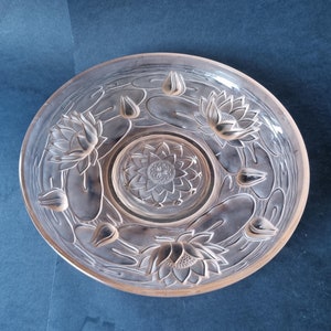May include: A clear, round glass serving dish with a floral lotus design. The dish features embossed lotus flowers and lily pads around the rim and centre. The dish is approximately 25 cm in diameter.
