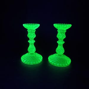 Puede incluir: Un par de candelabros de vidrio verde neón brillante con un borde superior festoneado, un tallo delgado con un patrón de espiral retorcida y una base circular ancha con detalles de remolinos radiantes, iluminados sobre un fondo oscuro.