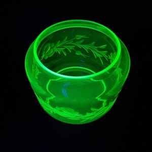 Op de afbeelding: Een heldergroene glazen kom met geëtste bloemmotieven. De kom heeft een ronde vorm en staat tegen een zwarte achtergrond. Het glas lijkt vintage, met delicate patronen rond de rand en de basis.