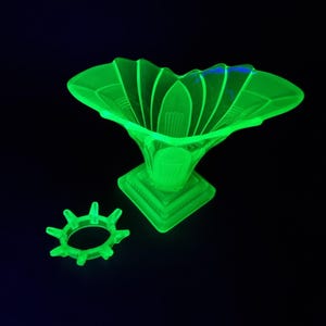 Könnte beinhalten: Eine leuchtend grüne Glasvase mit einem ausgestellten, blütenblattartigen Rand und einem quadratischen Sockel. Daneben ein passender grüner Ring mit spitzen Kanten. Die Objekte sind vor einem dunklen Hintergrund platziert.