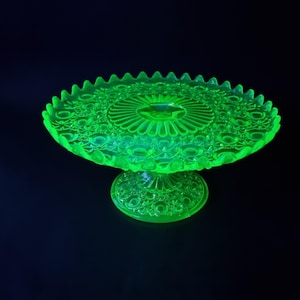 May include: A vibrant green glass cake stand with a detailed, textured pattern. The stand features a scalloped edge and a circular design on the base and top. The glass glows brightly against a dark background.