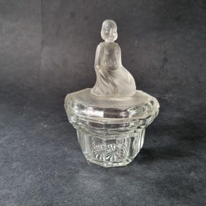 Könnte beinhalten: Eine transparente Glas-Schmuckschatulle mit einem Deckel aus mattiertem Glas, der eine sitzende Figur zeigt. Die Box hat ein geometrisches Design und einen dekorativen Sockel. Die Figur auf dem Deckel ist detailliert und verleiht dem Stück Eleganz.