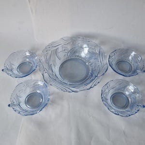 May include: A light blue glass serving set featuring a large bowl and four smaller bowls. The bowls have a decorative etched design and a rounded shape. The set is ideal for serving salads or snacks.