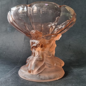 May include: A pink glass centerpiece bowl with a scalloped rim, supported by figures in a kneeling pose. The base has a detailed, etched design. The bowl is likely vintage or antique.