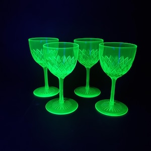 Op de afbeelding: Een set van vier oplichtende neongroene glazen op voet met een ruitpatroon op de kelk en een zonnestraalmotief op de ronde voet. De glazen stralen een helder fluorescerend licht uit tegen een donkere achtergrond, wat de textuur benadrukt.