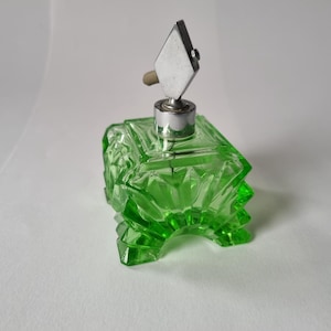 Puede incluir: Un frasco de perfume de vidrio verde con un diseño geométrico. El frasco tiene forma cuadrada con una base decorativa y una parte superior plateada con un acento en forma de diamante. El frasco está vacío.