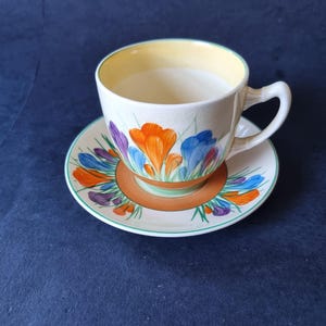 May include: A ceramic teacup and saucer set featuring a cream-colored base with a hand-painted floral pattern of orange, blue, and purple crocus flowers. The cup has a pale yellow interior rim, a curved handle, and a matching saucer with a brown band.