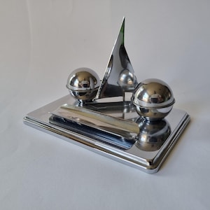 May include: A polished chrome desk accessory featuring a rectangular base with a central triangular sail-shaped element, flanked by two spherical containers. A long, narrow, curved tray is positioned in the front section of the metallic base.