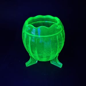 Op de afbeelding: Een kleine, ronde, doorschijnende groene glazen kom met een geschulpte rand en drie korte, stevige voetjes. Het buitenoppervlak van de kom is versierd met reliëf bloemmotieven en verticale ribbels, fel oplichtend tegen een donkere achtergrond.