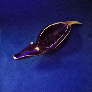 Puede incluir: Un plato de vidrio de color morado oscuro con forma de animal alargado, con un hocico puntiagudo y una cola. Los bordes del plato están delineados con un borde de vidrio fino de color claro, y dos pequeños acentos circulares claros cerca del hocico.
