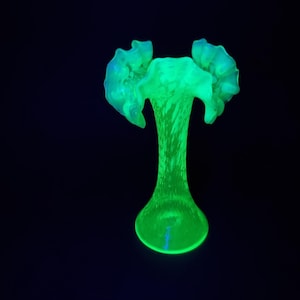 Può includere: Un vaso in vetro verde neon brillante caratterizzato da un collo sottile che si allarga in un bordo svasato e arricciato. La superficie presenta un motivo testurizzato e striato su tutto il corpo, emettendo una luce vivida su uno sfondo scuro.