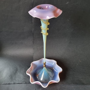 May include: A tall glass epergne featuring a wide, ruffled lavender top bowl and a matching ruffled base. The slender, pale blue stem is accented with a spiralling yellow glass coil and connects to the base with a small, round, metallic-toned bead.