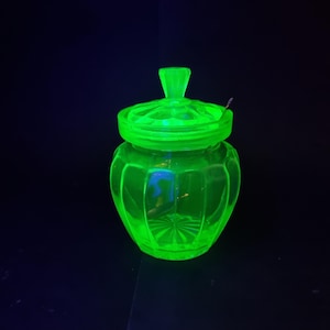 May include: A glowing green glass jar with a lid and a small spoon. The jar has a rounded shape with vertical ridges and a starburst pattern on the bottom. The lid has a decorative handle.