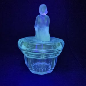 May include: A clear glass powder jar featuring a faceted base with a starburst pattern on the bottom and a lid topped with a frosted glass figurine of a kneeling person. The glass emits a bright neon green glow under ultraviolet light.