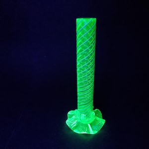 以下が含まれることがあります： 明るい緑色のガラスの花瓶。ベースはフレアで、円筒形の本体。薄い青色のラインのらせん模様があります。ベースはフリルとスカラップのデザイン。暗い背景にあります。