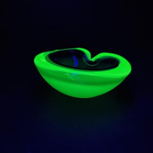 Puede incluir: Un cenicero verde neón con interior oscuro. El cenicero tiene una forma curva única y está sobre un fondo oscuro. El color verde vibrante es llamativo.