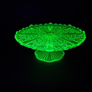 Puede incluir: Una brillante fuente para pasteles de vidrio verde con un diseño calado detallado. La fuente tiene una parte superior redonda y festoneada y una base de pedestal. El intrincado patrón y el color brillante crean un efecto visual llamativo sobre un fondo negro.