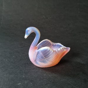 Puede incluir: Figurita decorativa de cisne de cristal en tonos rosa, azul y naranja. El cisne tiene un cuello curvado y alas detalladas, con un acabado suave e iridiscente. La figura es un objeto decorativo.