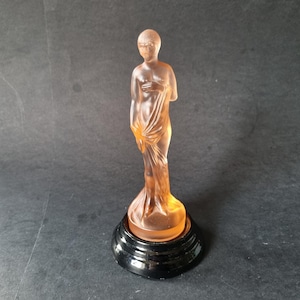 Puede incluir: Una figura art déco de color melocotón de una mujer drapeada de pie sobre una base negra escalonada. La estatua es translúcida, permitiendo el paso de la luz, y está sobre un fondo oscuro.