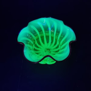 Puede incluir: Un cuenco de cristal verde brillante con un borde festoneado y ondulado y un patrón de nervaduras en espiral. El cuenco emite una luz verde neón brillante contra un fondo oscuro, resaltando la estructura de cristal translúcido y el diseño curvo.