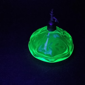 May include: A vibrant green glass perfume bottle with a decorative stopper. The bottle is round with etched floral patterns. The stopper is dark and ornate. The bottle sits on a dark background.