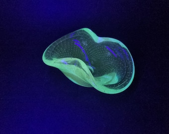 Può includere: Una ciotola in vetro traslucido e ondulato caratterizzata da un distinto motivo a bolle in tutta la sua struttura. Il vetro emette una vivida luce verde neon brillante, che indica una reazione alla luce ultravioletta, su uno sfondo scuro e uniforme.