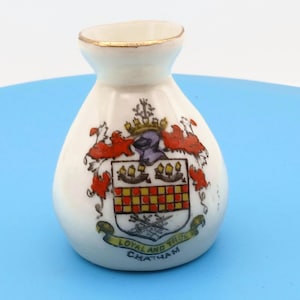 May include: A small white ceramic vase with a gold-trimmed rim, featuring a colorful heraldic crest on the front. The crest includes a crown, red and white mantling, and a shield with a checkered pattern. Below the shield is a banner with the text "LOYAL AND TRUE CHATHAM".
