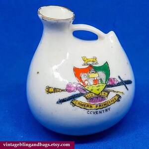 May include: A small white porcelain vase with a handle. The vase has a colorful crest with a lion, elephant, and swords. The crest includes the text "Camera Principis" and "Coventry".