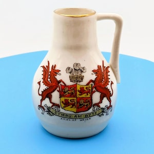 May include: A small, white ceramic pitcher with a handle and gold trim. The pitcher features a detailed coat of arms with red dragons, a crest, and the text "CYMRU AM BYTH ARMS OF WALES".