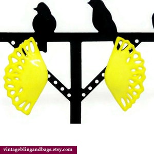 May include: A pair of yellow plastic earrings shaped like a fan with a scalloped edge. The earrings are displayed on a black bird-shaped earring stand.