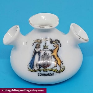 May include: White ceramic Edinburgh souvenir with three spouts and a central crest. The crest features a woman, a golden deer, and a castle, with the word "Edinburgh" below. The item has a vintage appearance.