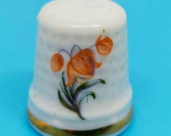 Peut inclure: Dé à coudre en porcelaine blanche avec un motif floral. Le dé à coudre a une bordure dorée autour de la base et présente des fleurs orange avec des feuilles vertes.