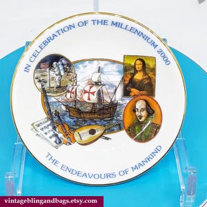 Puede incluir: Un plato de cerámica blanco con borde dorado presenta un diseño colorido que celebra el milenio. El diseño incluye imágenes de un barco, un laúd, un astronauta, William Shakespeare y la Mona Lisa. El texto en el plato dice "In Celebration of the Millennium 2000 The Endeavours of Mankind".