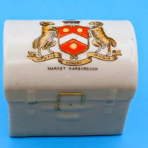 May include: A white ceramic trinket box shaped like a chest, featuring a colorful heraldic crest with two rams, a red and white shield, and a gold banner with the text HOSTIS HONORI INVIDIA and the words MARKET HARBOROUGH printed below.
