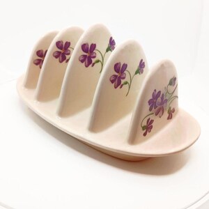 May include: A vintage ceramic toast rack in a soft cream color, featuring five arched slots. Each slot is adorned with a delicate purple floral design. The rack has an oval base.