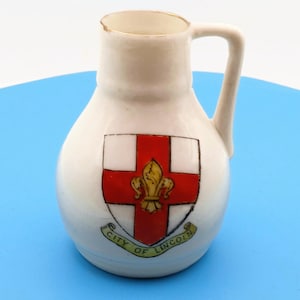 May include: A small, white ceramic pitcher with a handle and a gold rim. The pitcher features a red cross with a gold fleur-de-lis in the center, and the words "CITY OF LINCOLN" in a banner below the cross.