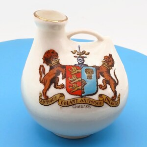 Puede incluir: Jarra de cerámica blanca con asa y pico, con un escudo con dos leones marrones, un escudo rojo y azul, y las palabras "ANTIQUAM COLANT ANTIQUAM DILRUM CHESTER" en una pancarta. El pico tiene un borde dorado.