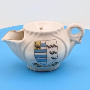 Op de afbeelding: Een witte keramische creamer met een handvat en tuit. De creamer heeft een wapen met een vogel en blauwe strepen. Het woord "Bognor" staat op de bodem van de creamer.