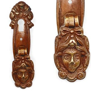 Op de afbeelding: Twee antieke bronzen deurgrepen met een gedetailleerd gezichtsontwerp. De grepen hebben een sleutelgat en een decoratief gezicht met een strik en sierlijke details. Het brons heeft een warme, verouderde patina en het gezicht is geaccentueerd met gouden accenten.